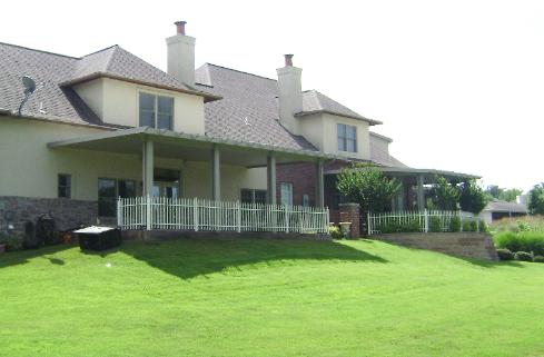 Large house with a covered patio and white railing, overlooking a bright green lawn under a cloudy sky