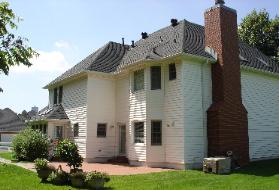 Two-story beige house with a brick chimney, dark shingled roof, and green lawn in daylight