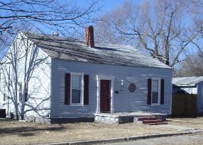 Small gray house with maroon shutters and a red bench in front, seen from the street.