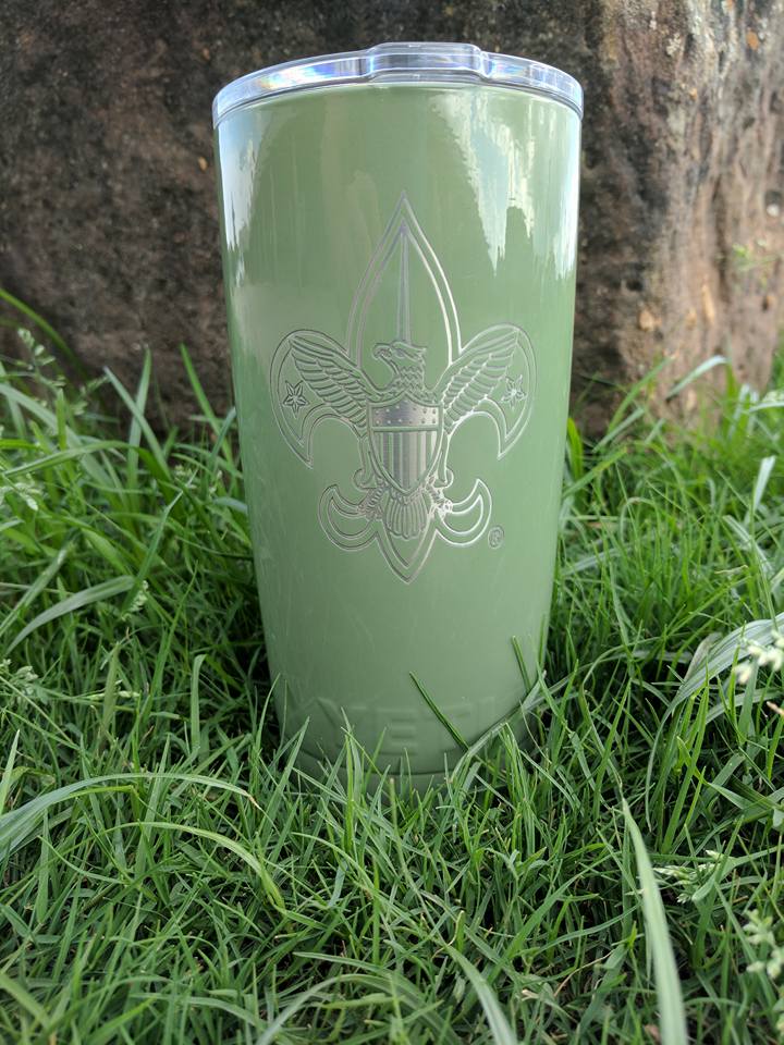 Green insulated tumbler with engraved fleur-de-lis design, set on grass in front of a rock.