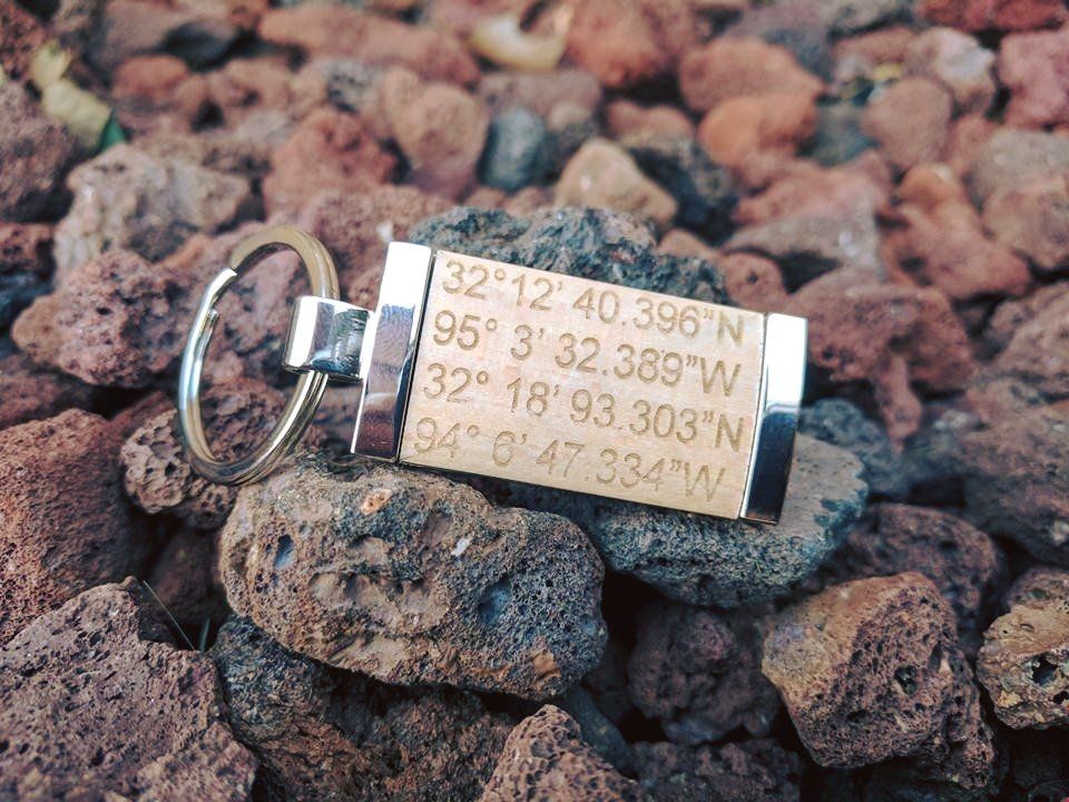 Keychain with engraved GPS coordinates on a bed of reddish-brown rocks.