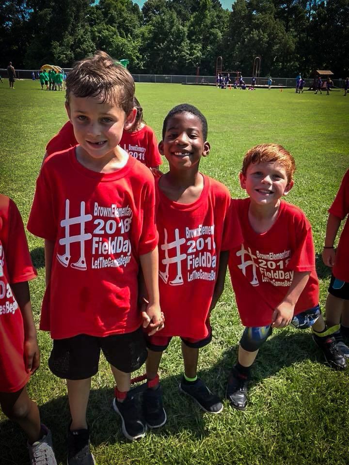 Three smiling children in red shirts on a grassy field. Shirts say 