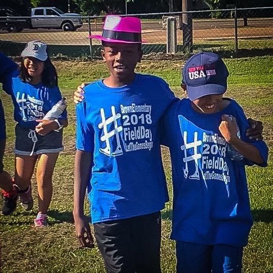 Two children in blue shirts and hats pose on a grassy field; a third child is in the background.