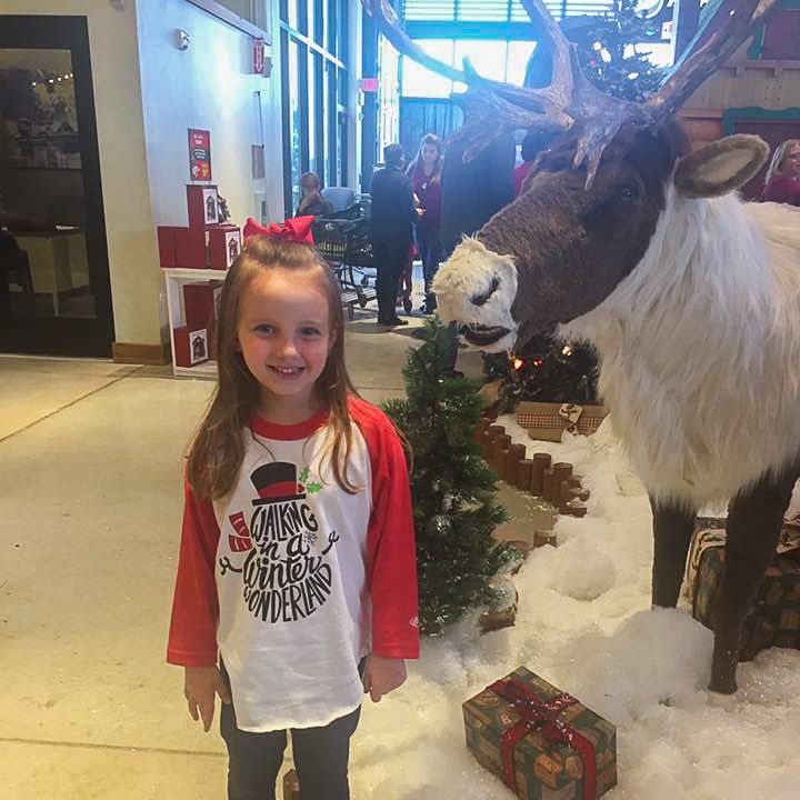 Young girl smiles in front of a reindeer display with a Christmas-themed shirt; indoors.