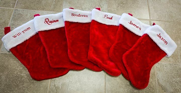 Six red Christmas stockings with white cuffs, each embroidered with a different name.