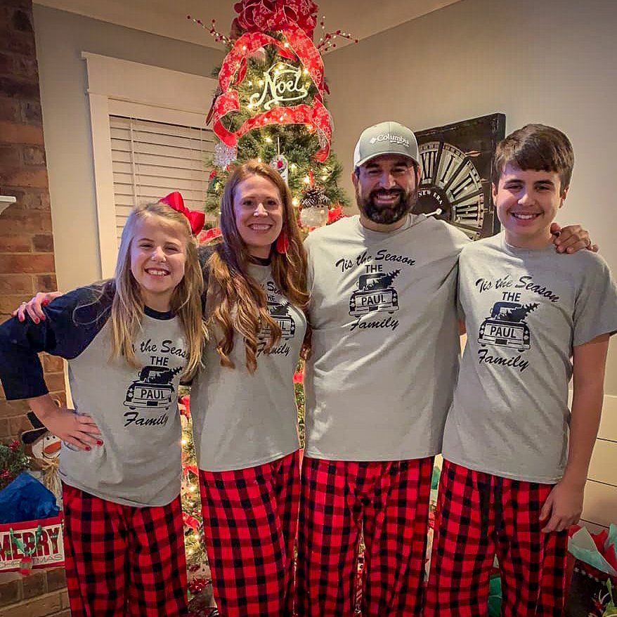 Family wearing matching pajamas stands in front of a Christmas tree and fireplace.