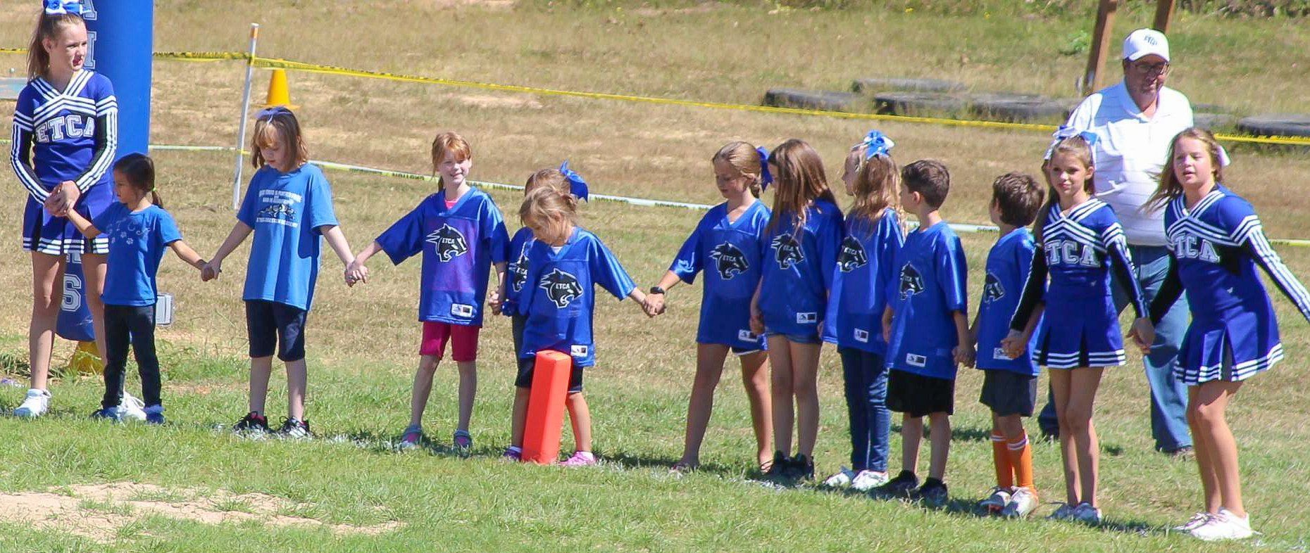 Children and cheerleaders hold hands on a grassy field, all wearing blue. A man in a white shirt stands behind them.