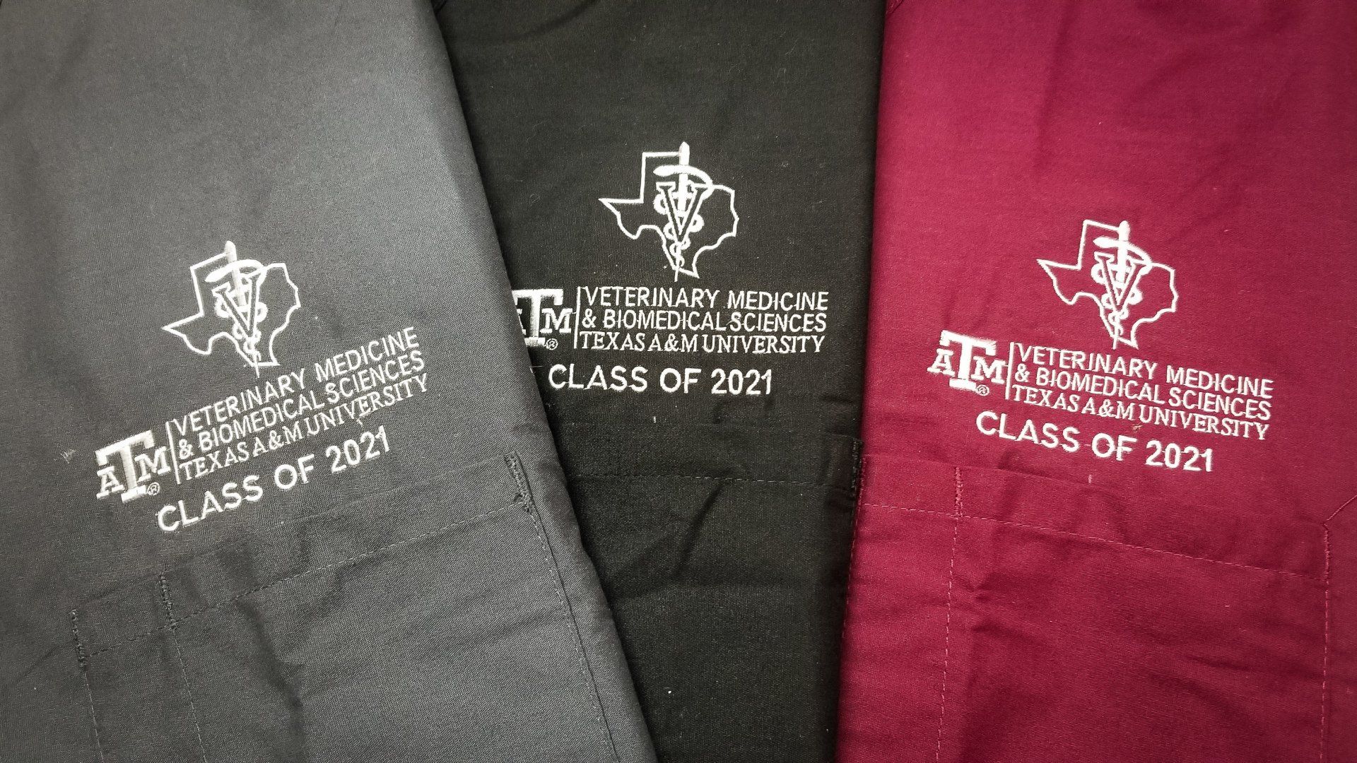 Three veterinary scrubs: gray, black, and maroon. Each has a white Texas A&M logo and 