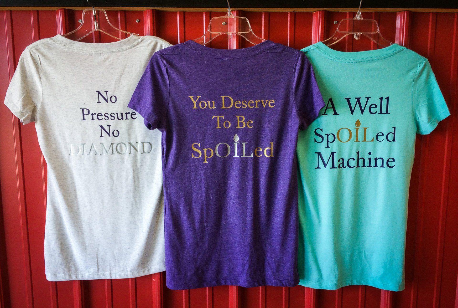 Three t-shirts hanging: white, purple, and teal, each with text on the back.