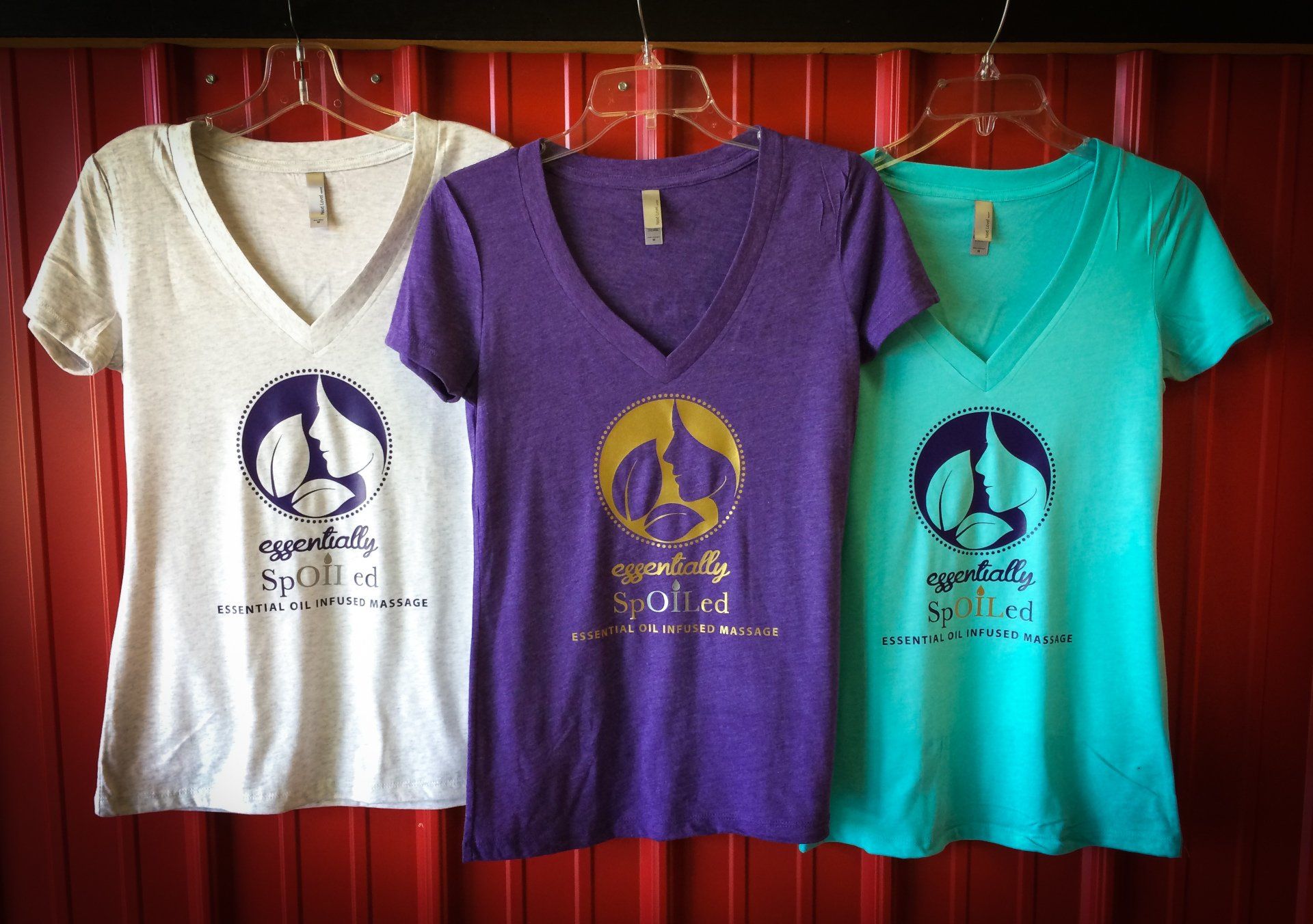 Three v-neck t-shirts in white, purple, and teal with a logo and text hanging in front of a red wall.