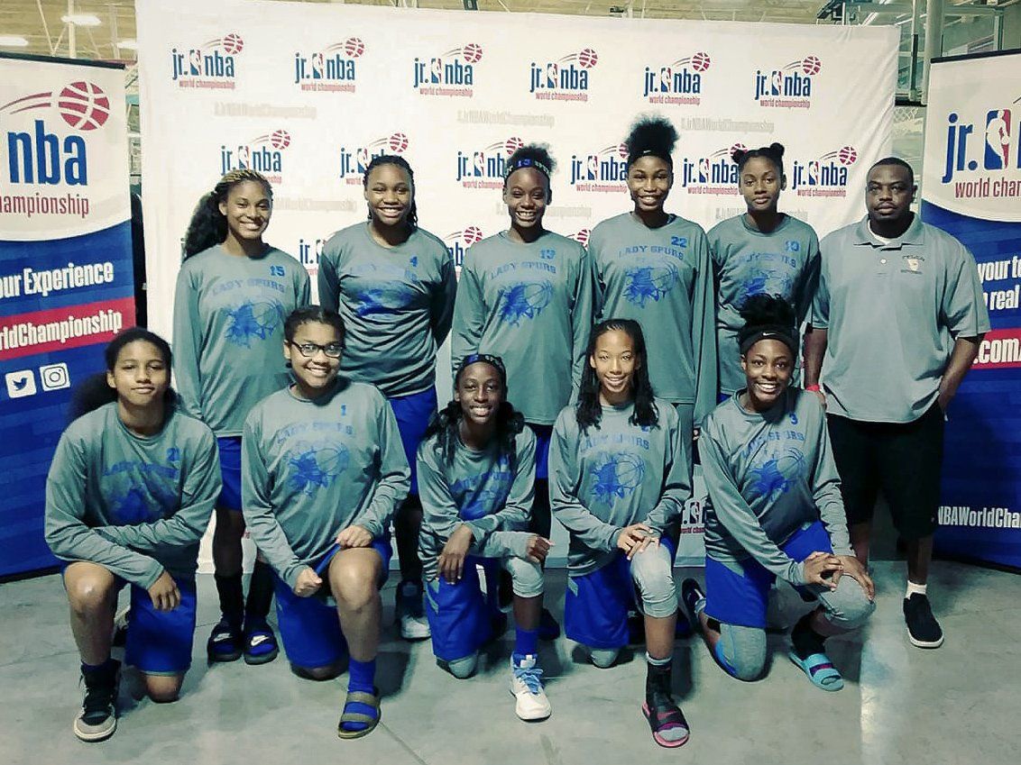 Basketball team poses in gray shirts with blue accents, in front of a banner.