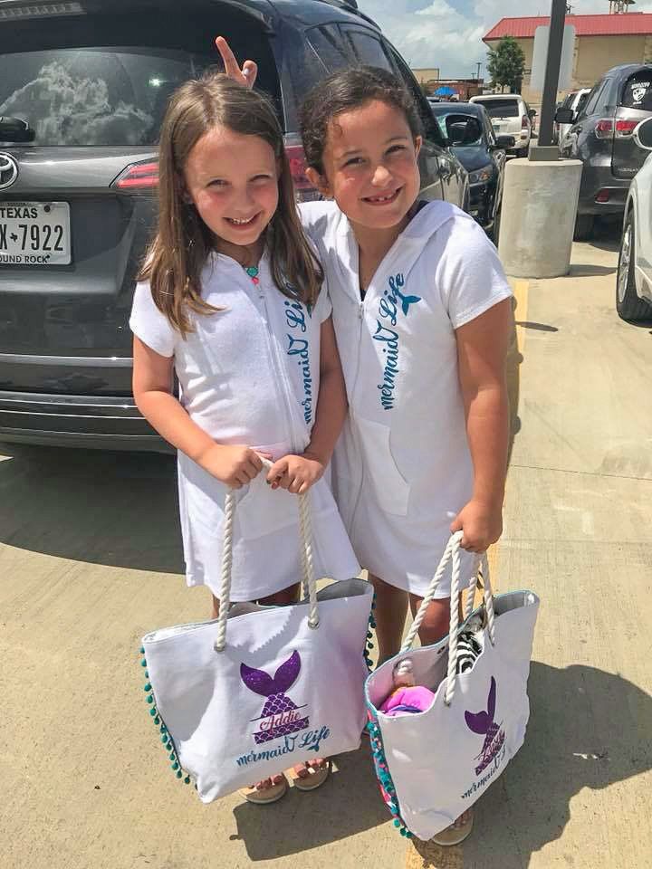 Two girls in white hooded dresses holding tote bags, posing with a car in a parking lot.