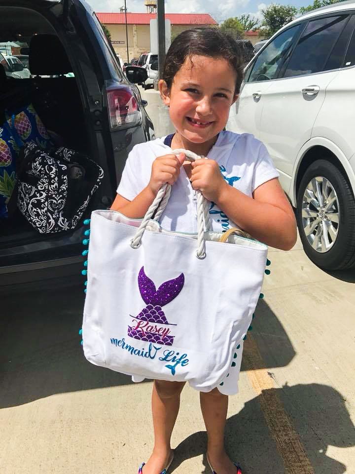 Girl holding a white tote bag with a purple mermaid tail design; smiling, outdoors near a car.