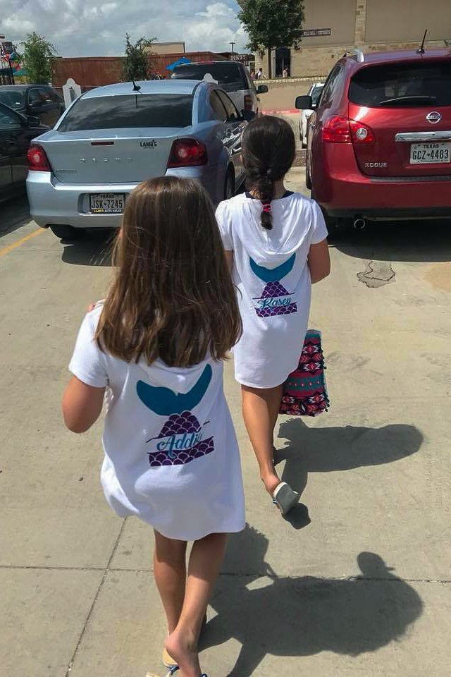 Two girls in white dresses with mermaid tail logos walk toward a parking lot.