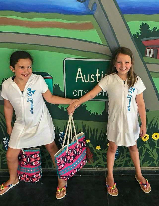 Two girls in white dresses holding patterned bags, posing in front of an Austin mural.