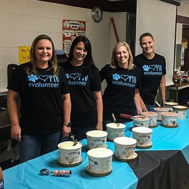 Four women in matching shirts serve ice cream from a table.