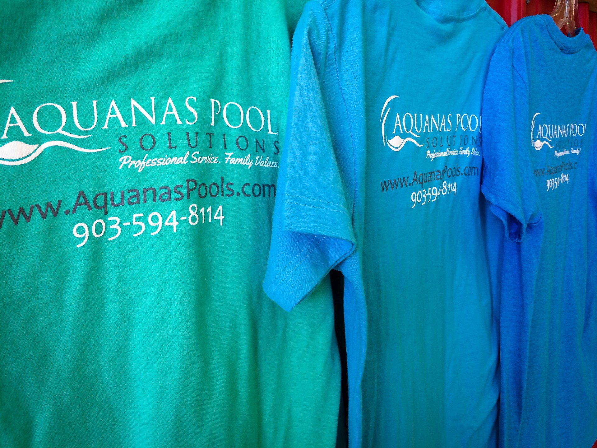 Three Aquanas Pool Solutions t-shirts in shades of blue and green, displayed vertically. Each has a logo and contact info.