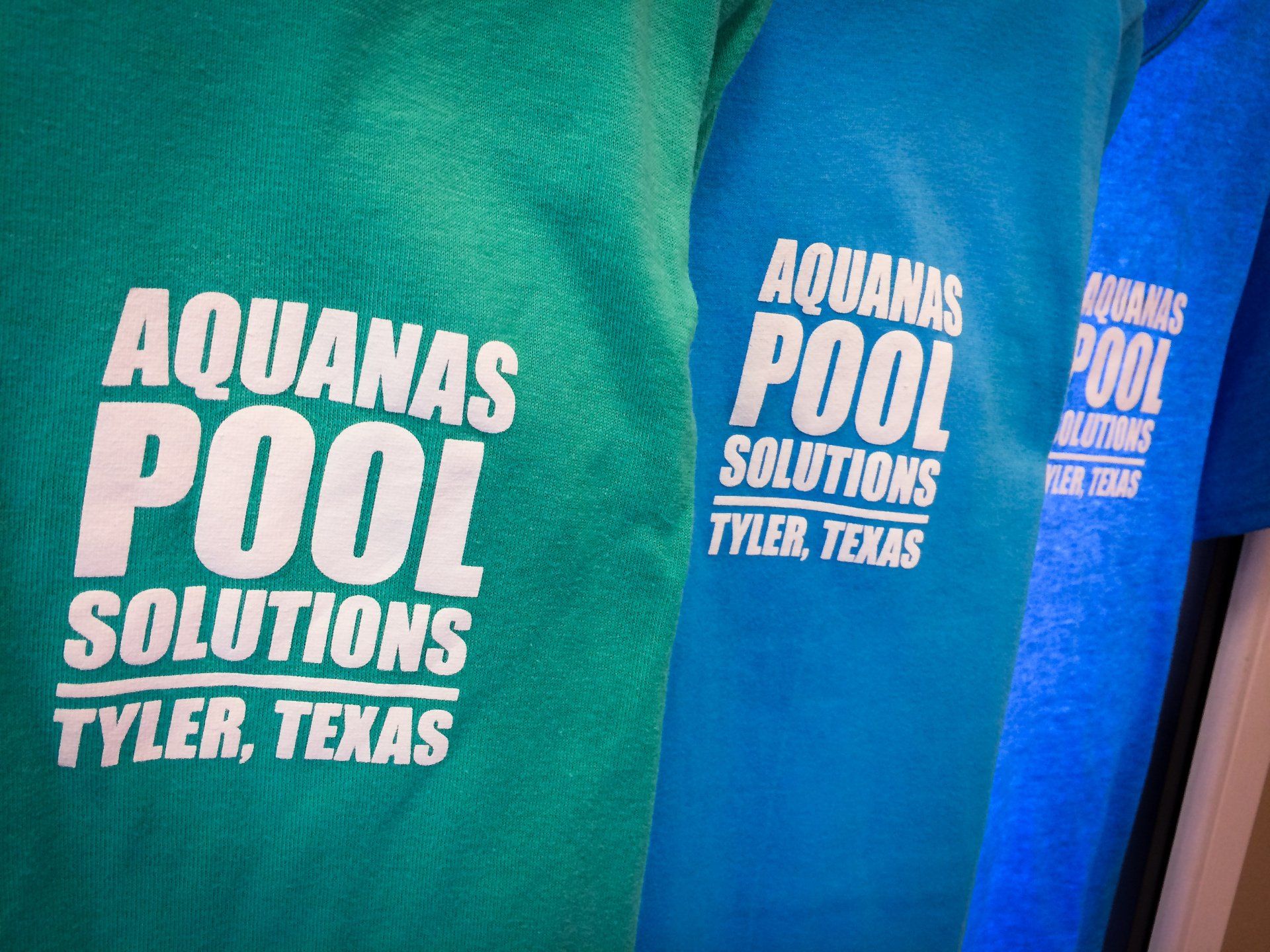 Close-up of three shirts in teal, turquoise, and blue with the text 