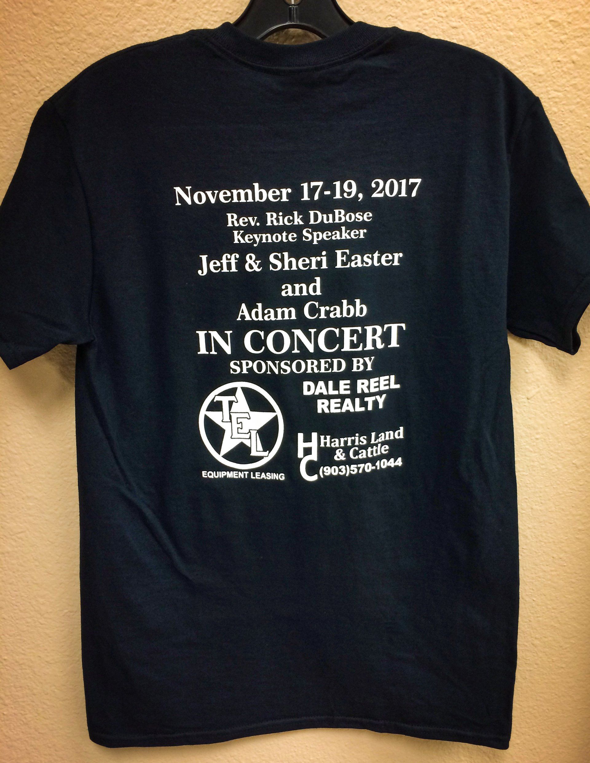 Black t-shirt with white text. It lists the date and names of performers and a real estate sponsor.