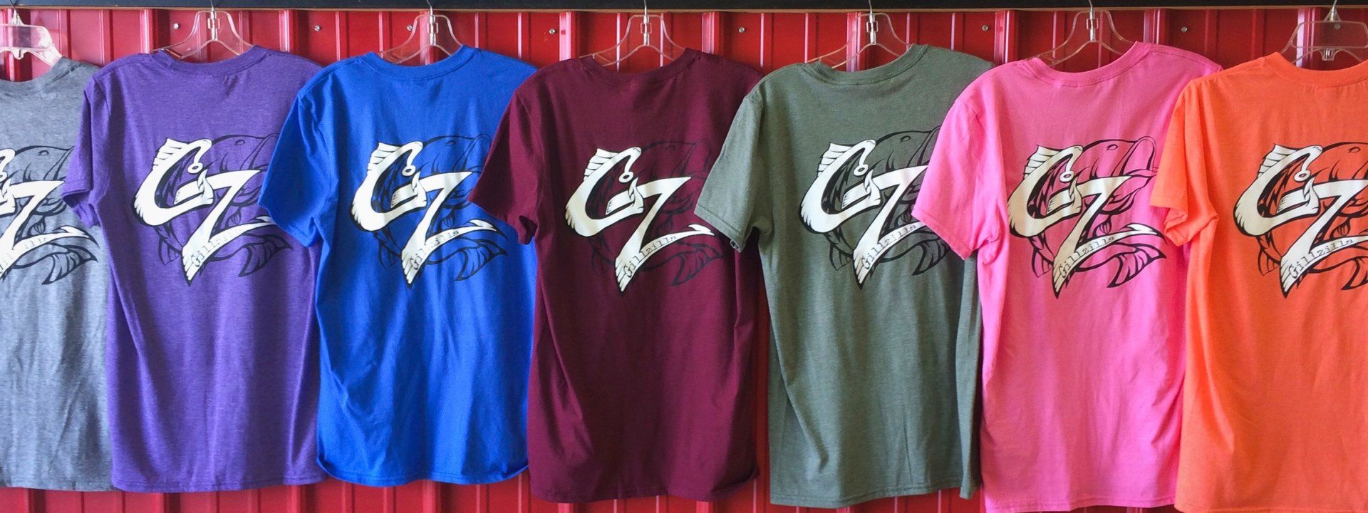 Row of colored t-shirts with white logo on them, hanging on a red background.