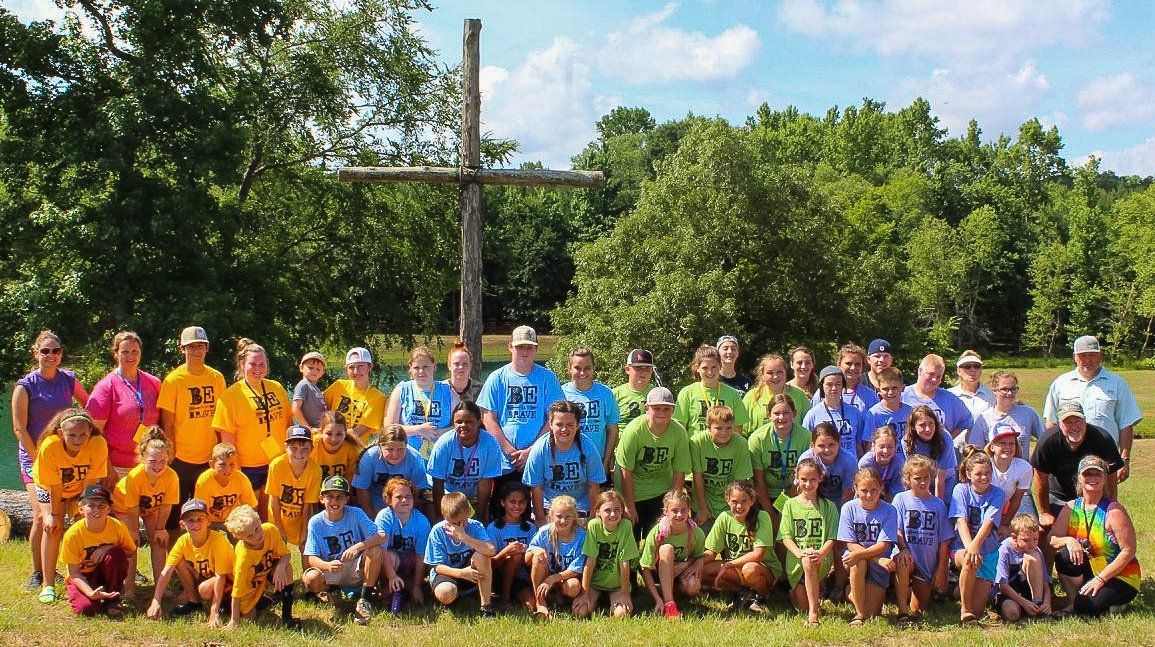 Group of people in colorful shirts pose in front of a large cross.