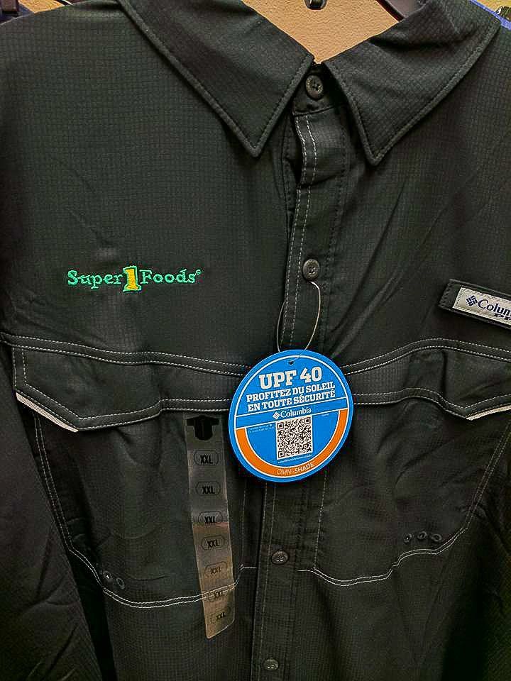 Black Columbia button-up shirt with Super Foods logo and hang tag, hanging.