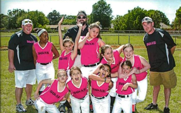 Softball team in pink and white uniforms with coaches, posing on a field.