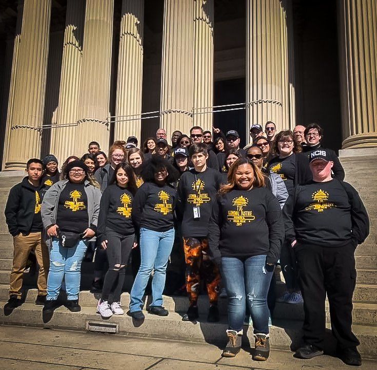 Group of people in black shirts with gold design, posed on steps in front of a building with columns.