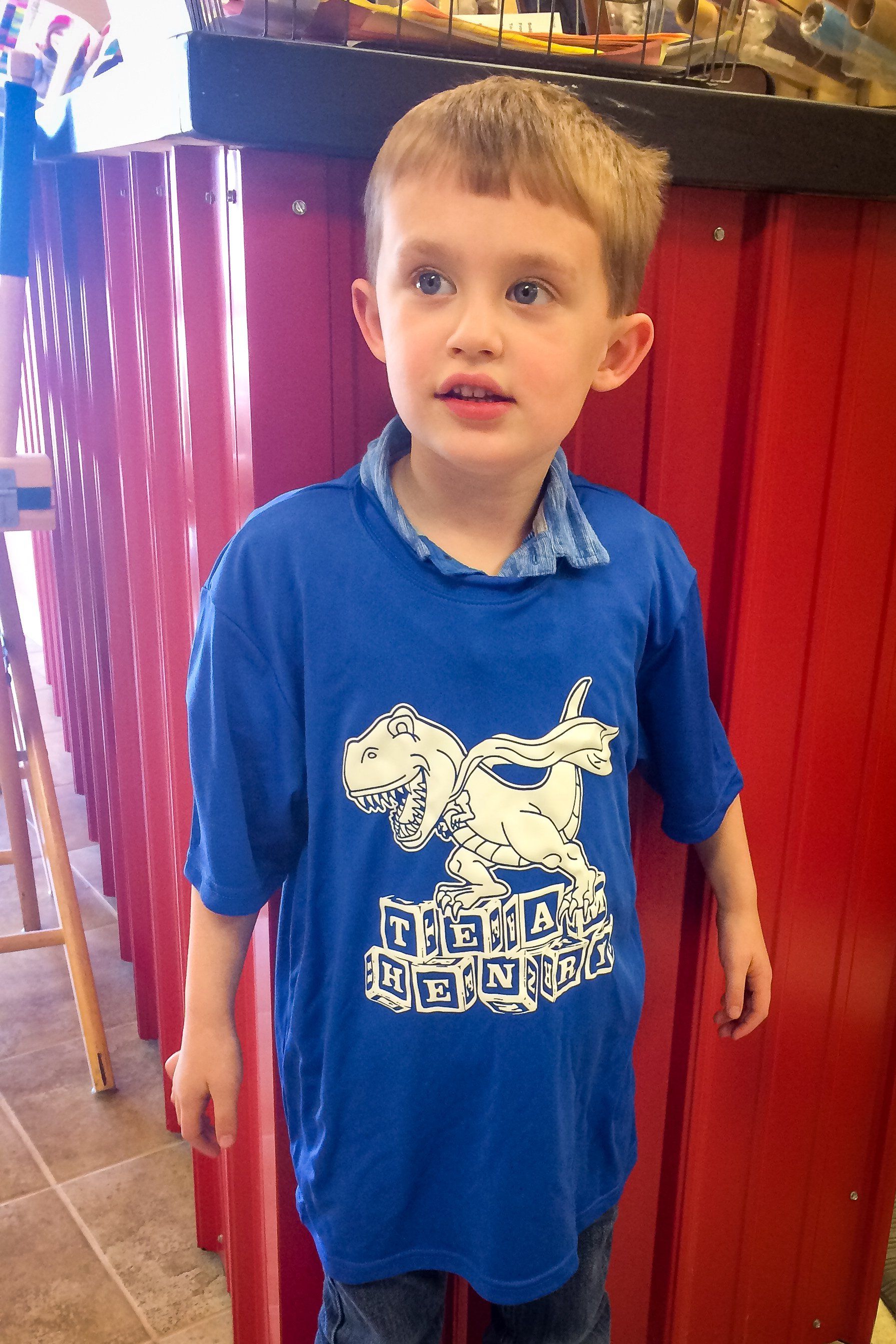 Boy in blue dinosaur shirt standing next to a red structure.