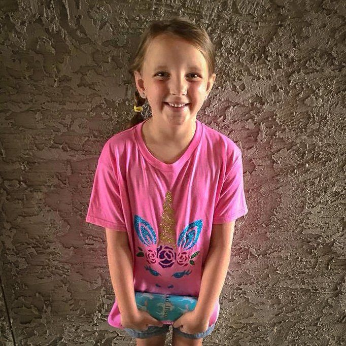 Girl with pigtails in pink unicorn shirt smiles, holding a blue item, against textured wall.