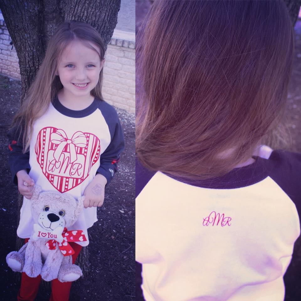 Girl smiling, holding stuffed dog. White shirt with red heart design. Back of shirt with embroidered initials.