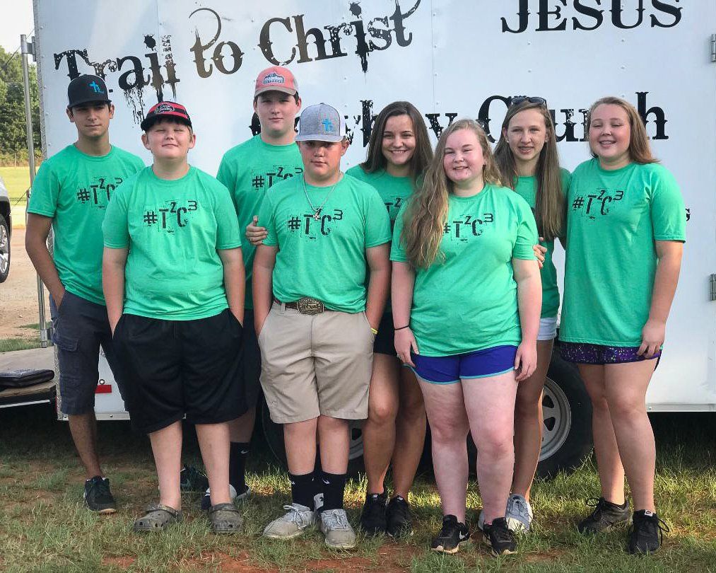 Group of people in green shirts, in front of a trailer, which reads “Trail to Christ.”