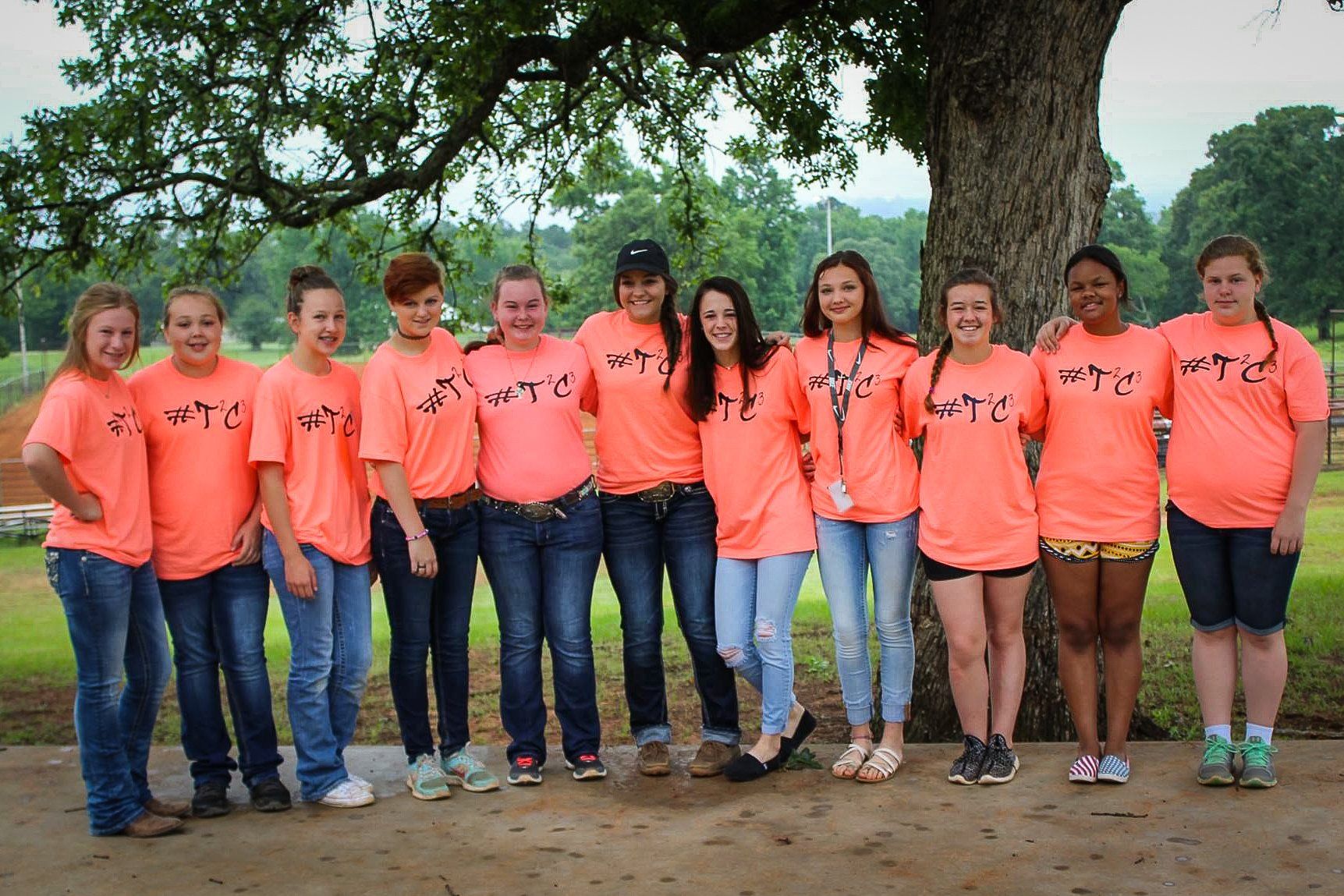 Group of people wearing orange shirts and jeans standing by a tree.