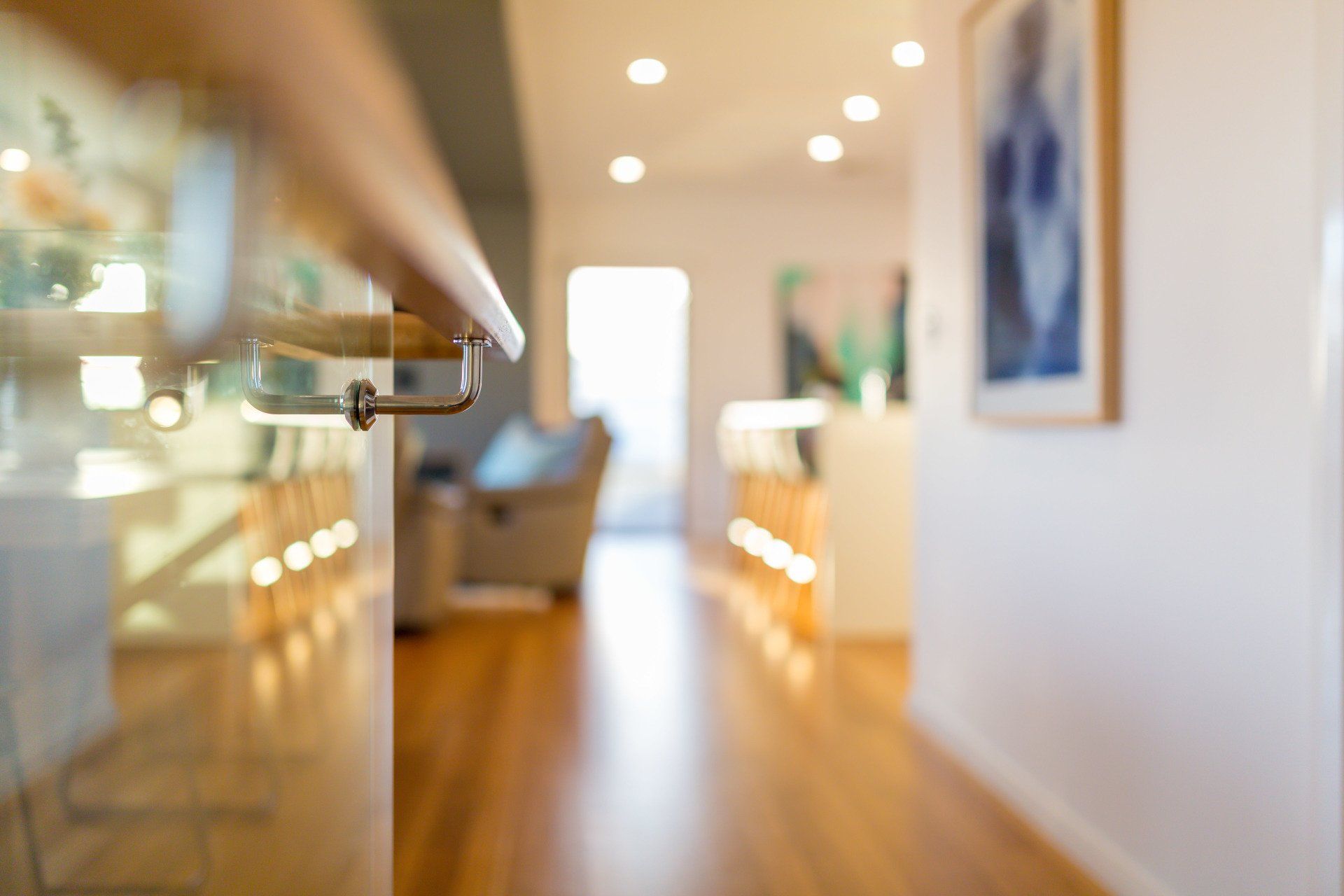 Hallway with wooden floor, open glass door with handrail, blurred background with art, and light fixtures.