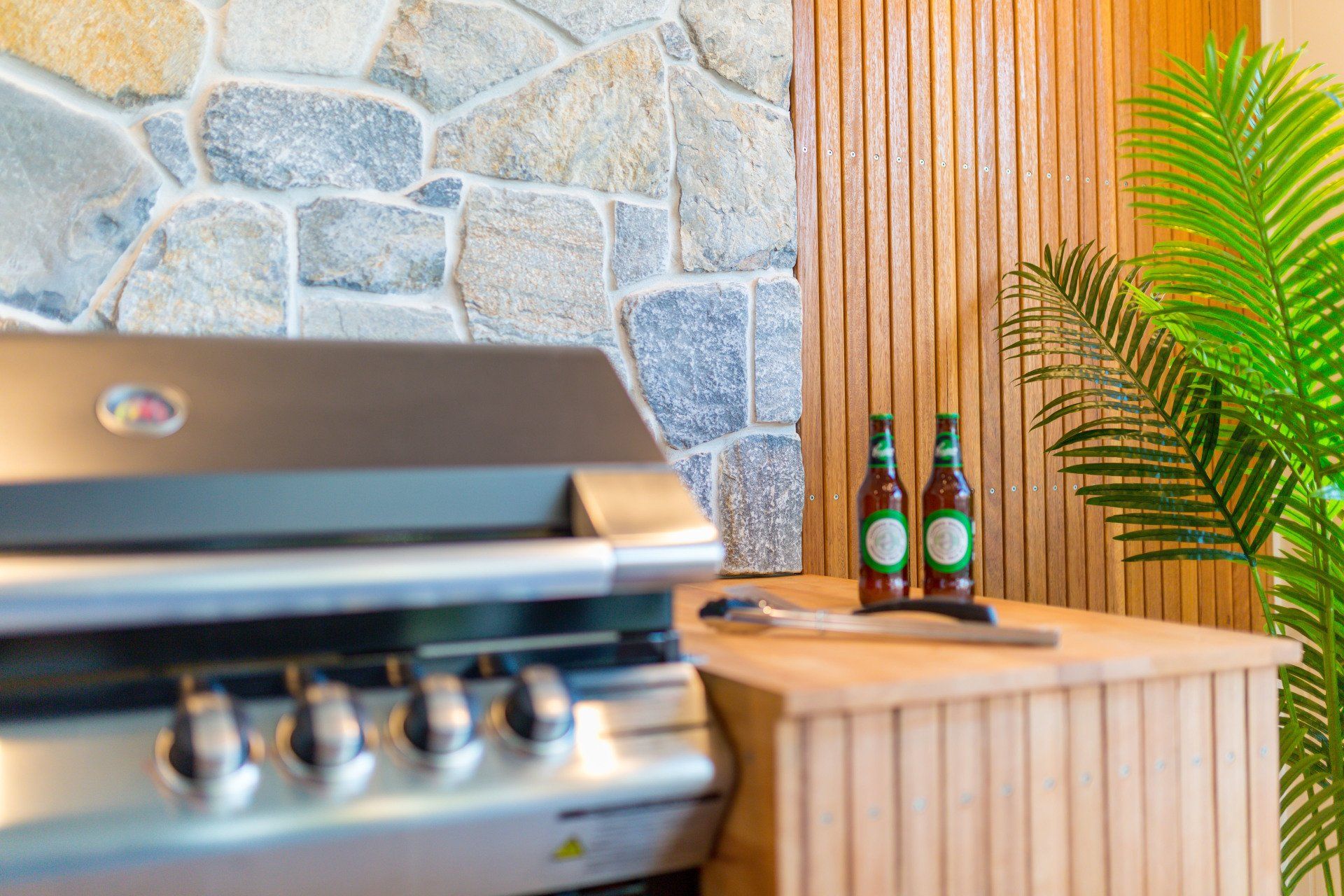 Stainless steel grill, beer bottles, and stone wall on a wooden counter, with a palm leaf.