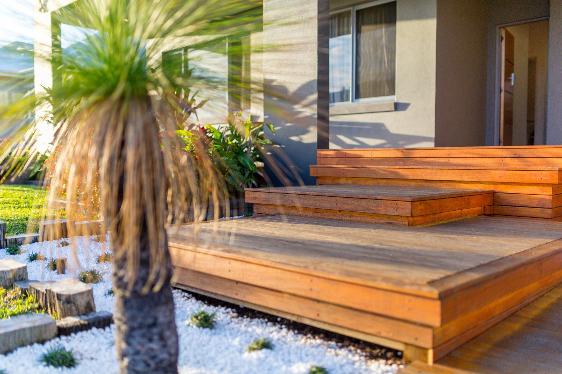 Wooden deck and steps with a gray house, a grass tree, and a white gravel garden.