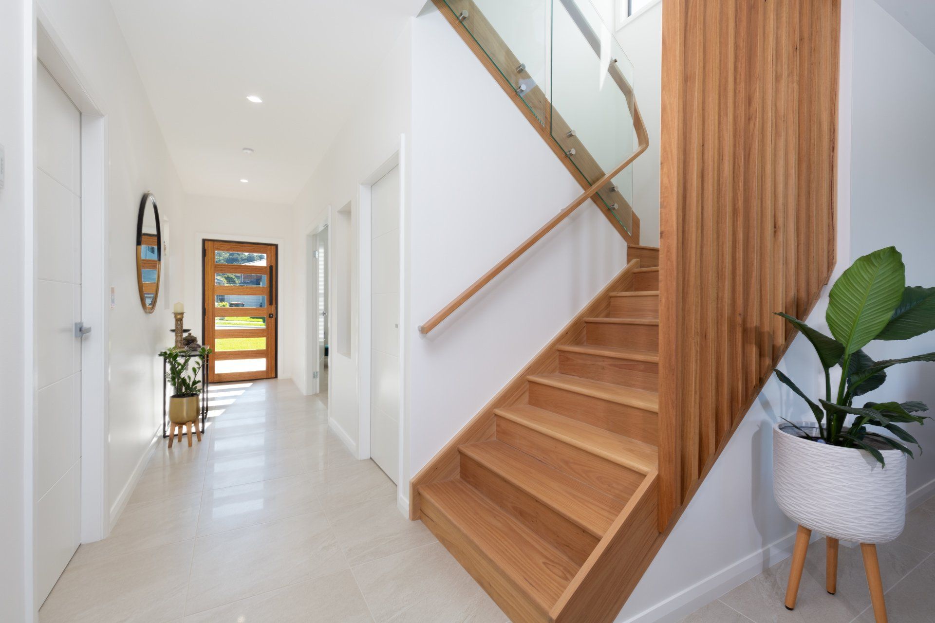 Hallway with wooden staircase and front door, white walls, natural light, potted plant.