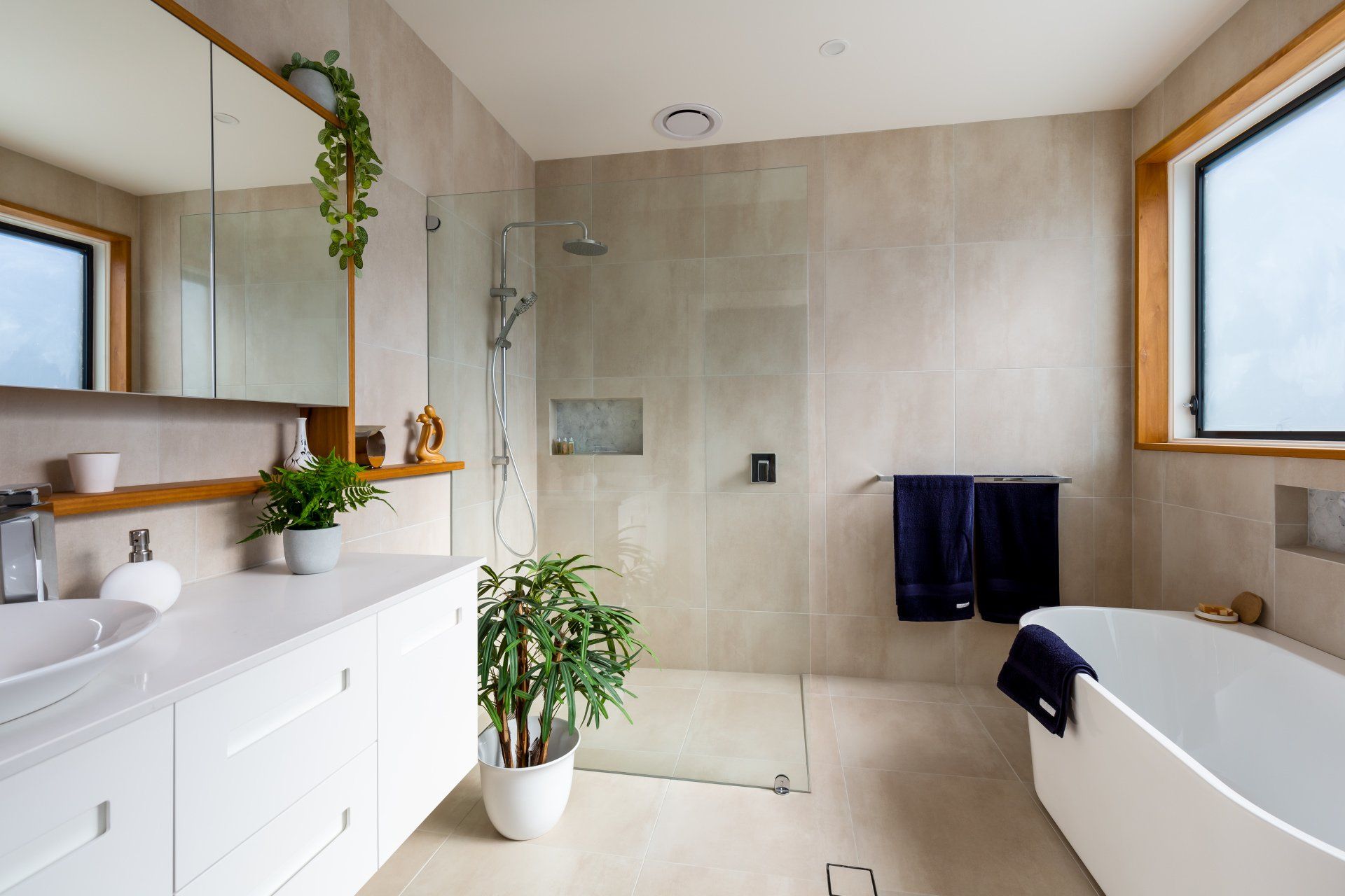Modern bathroom with a white vanity, walk-in shower, and a freestanding tub. Soft beige walls, wood trim, and greenery.