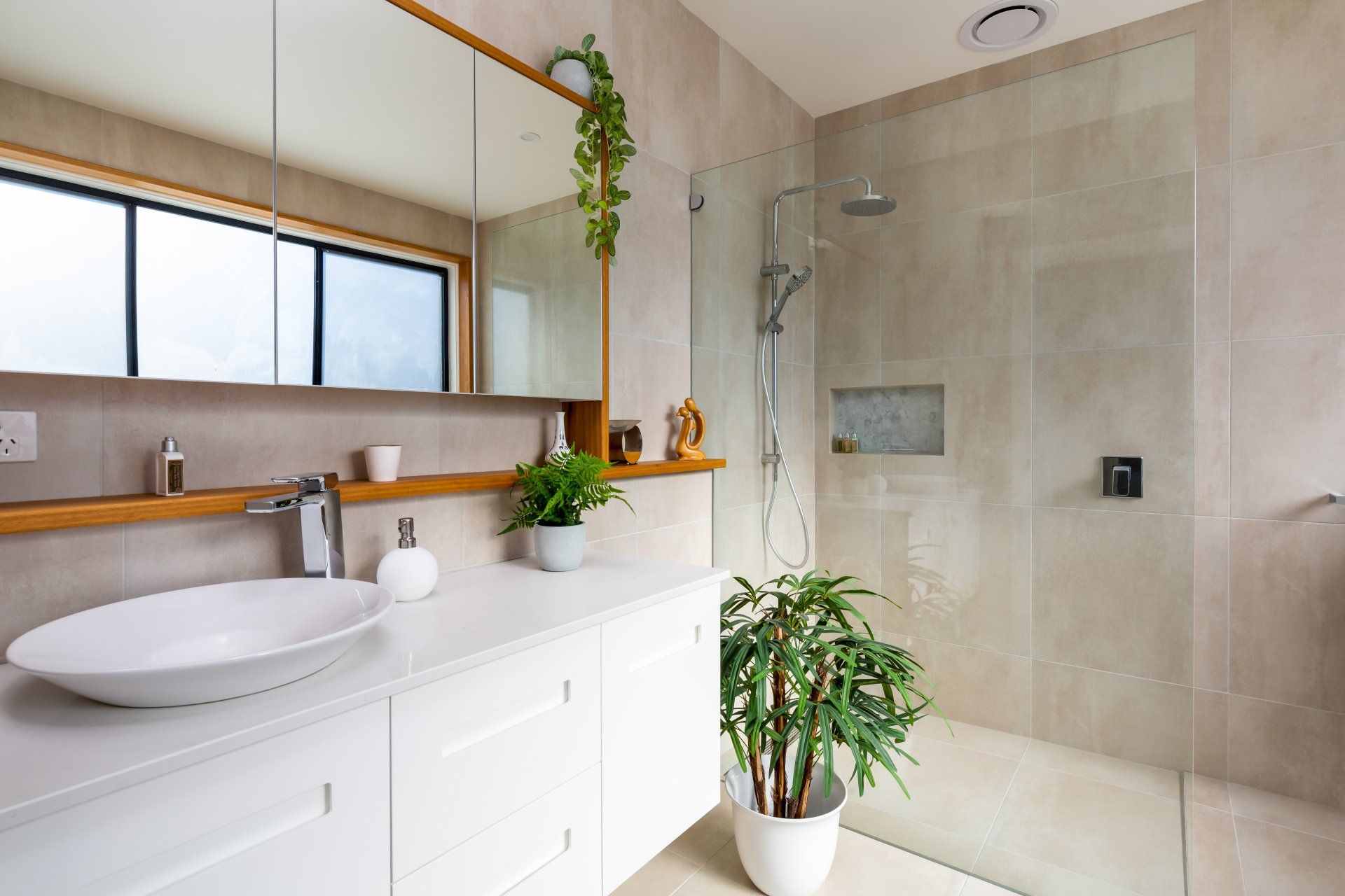 Modern bathroom with white vanity, glass shower, beige tile, and plants.