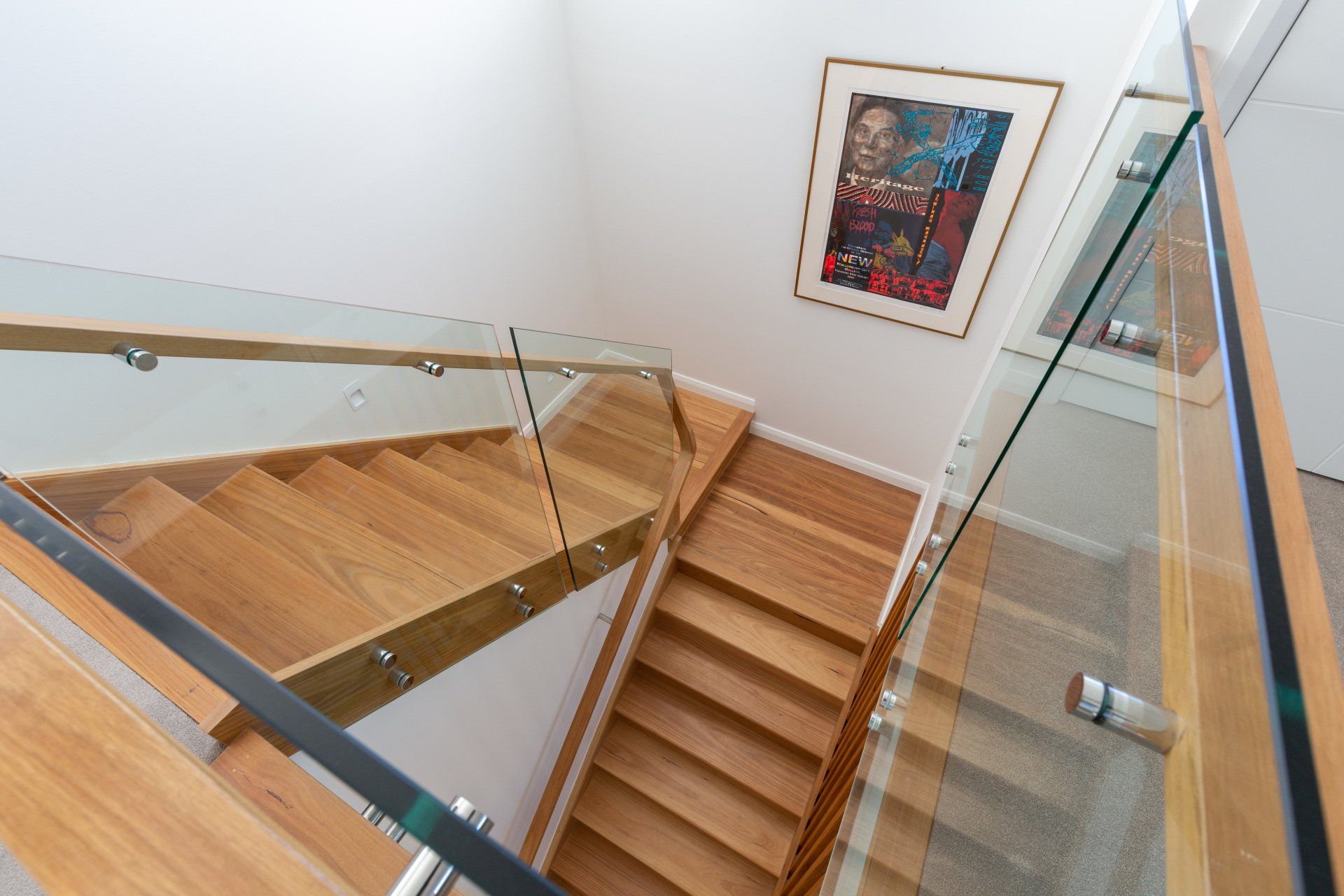 Wooden staircase with glass railings, ascending to a landing. A framed picture hangs on the wall.