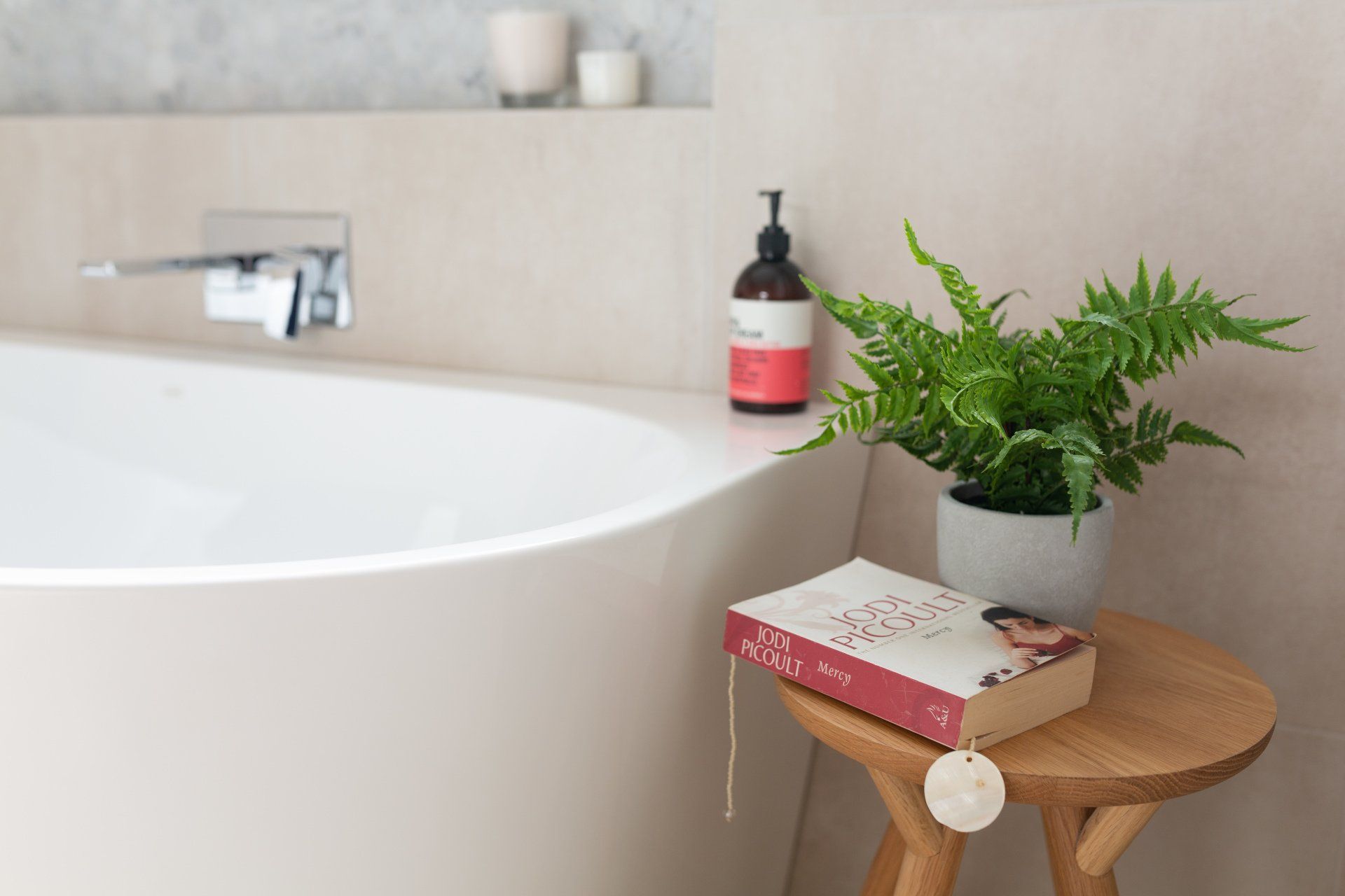 White bathtub with a book, potted fern, and soap on a small wooden table in a neutral-toned bathroom.