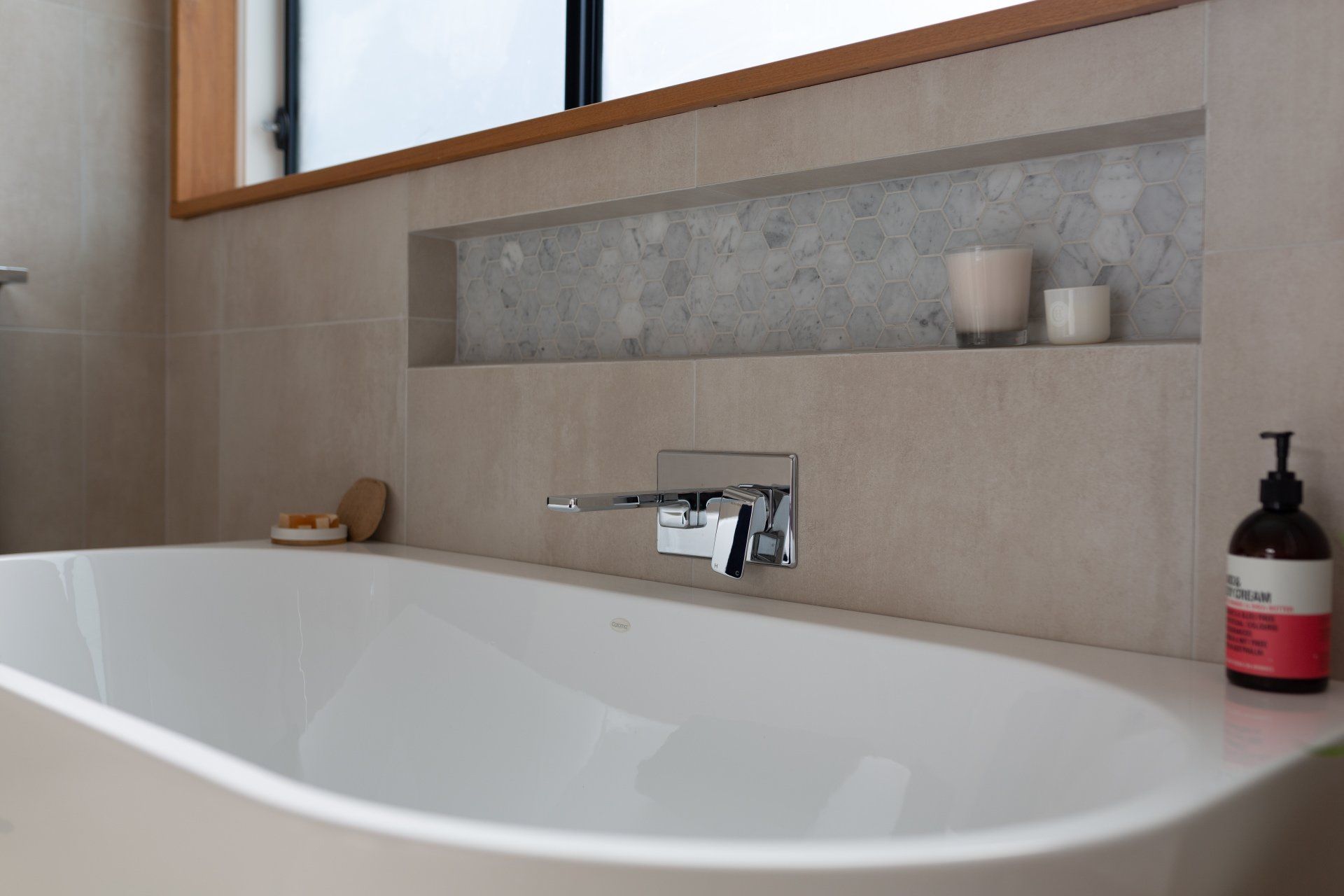 A white bathtub with chrome faucet, recessed shelf with candles, beige walls, and wood-framed window.