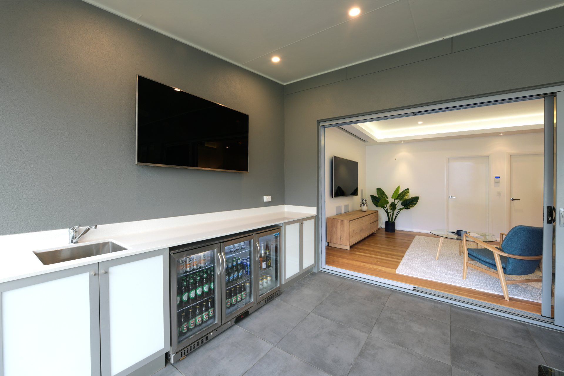A modern bar area with a TV, cooler, and sink.  Doorway leads to a living room with a TV, plant, and chair.