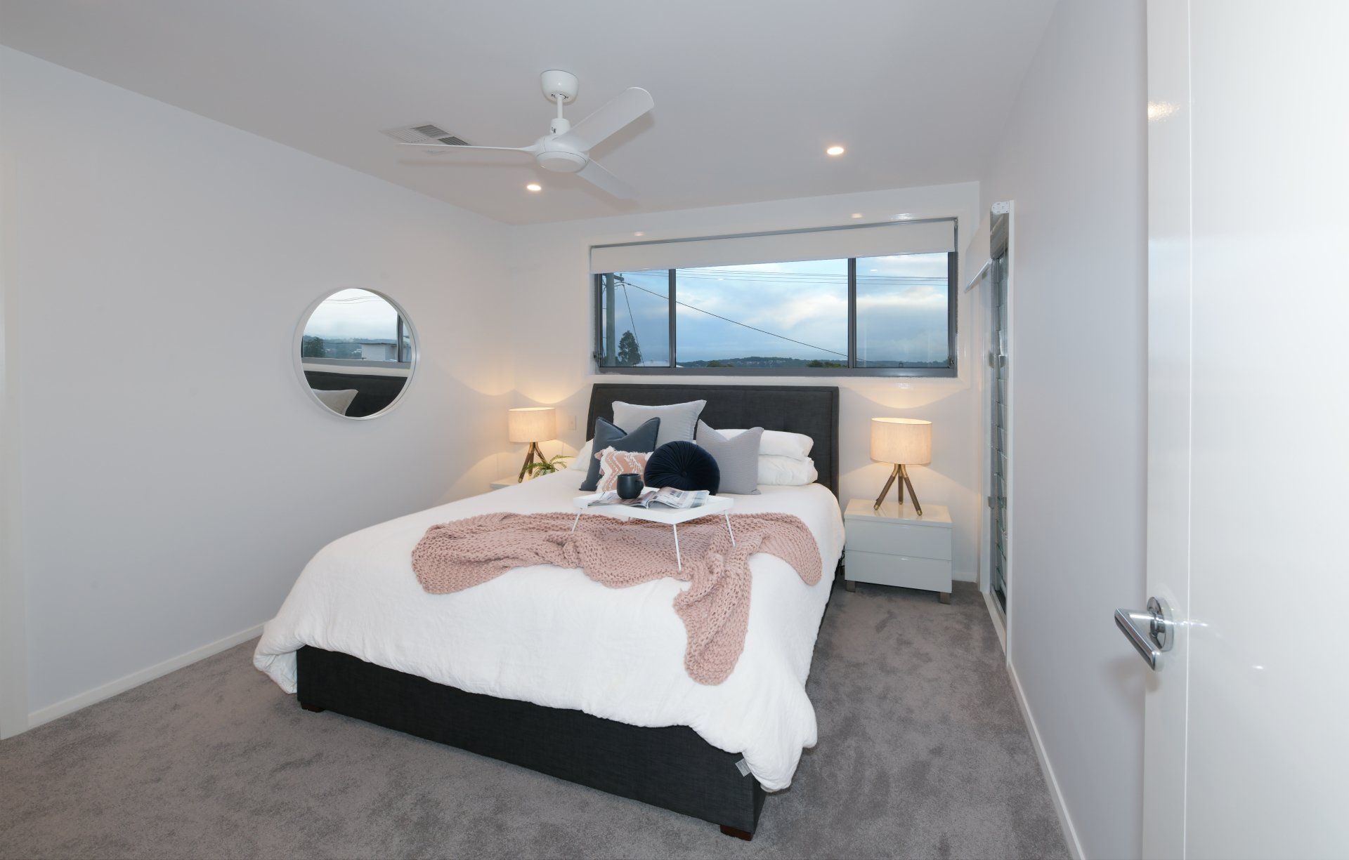 Bedroom with white walls, gray carpet, bed with white and pink bedding, and a round mirror.