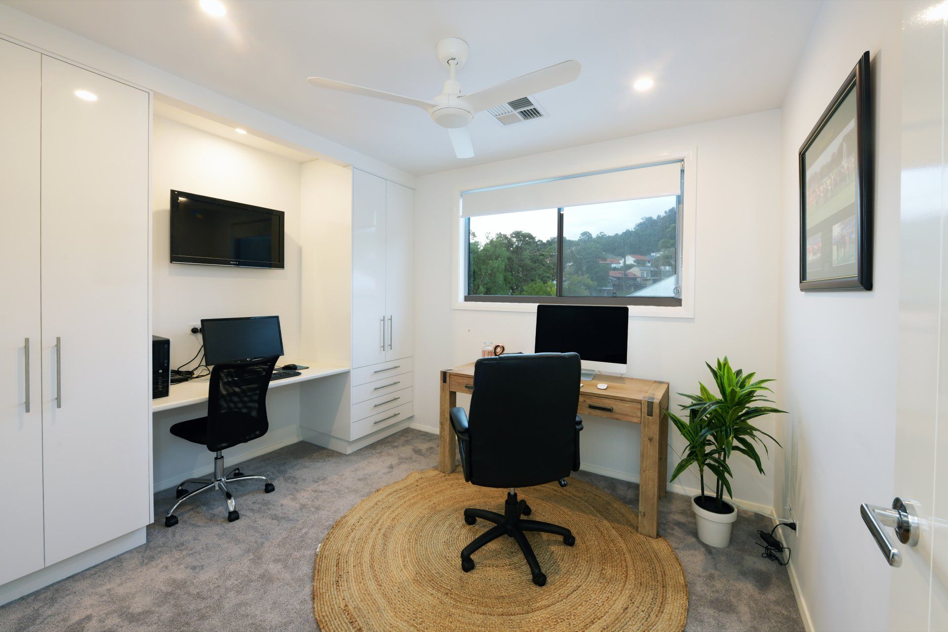Home office with two desks, built-in white cabinets, a rug, a plant, and a window.