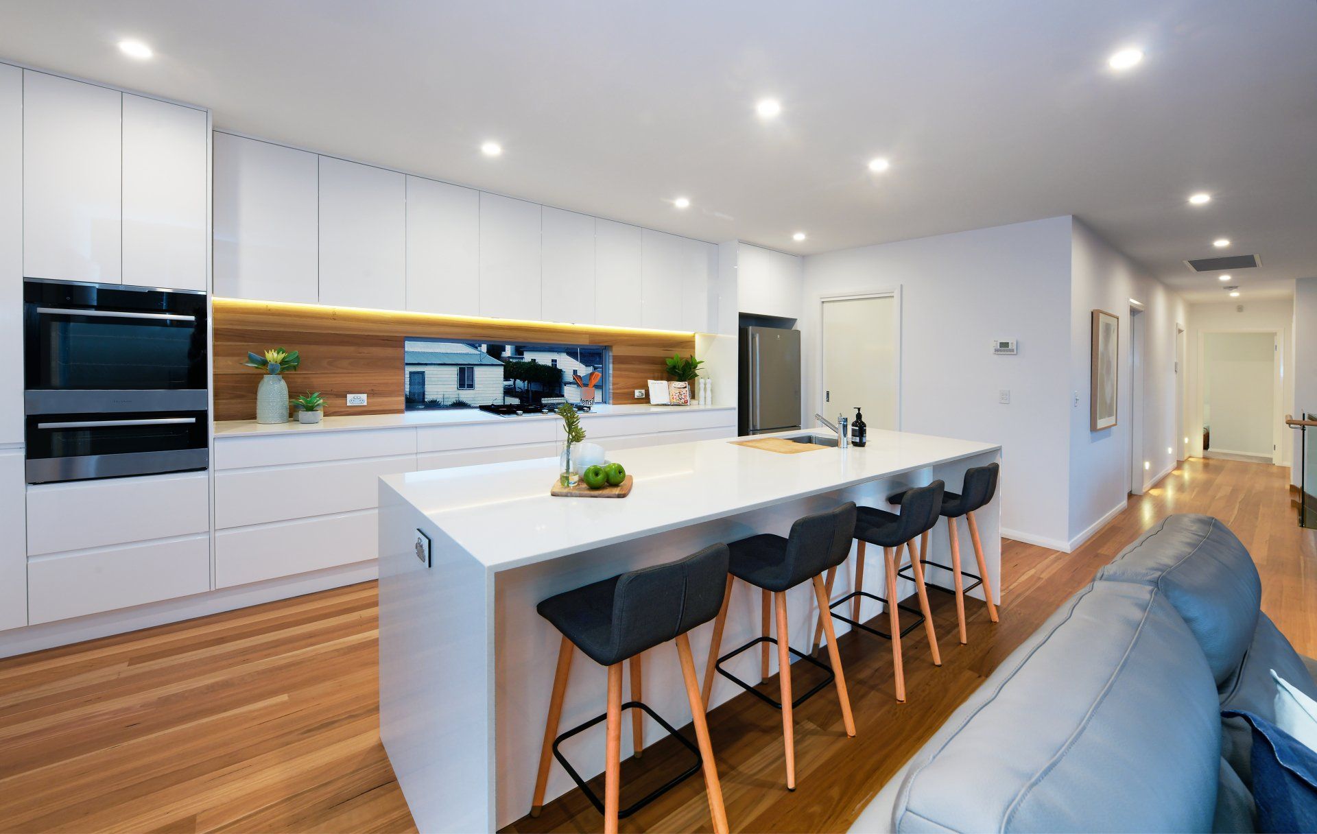 Modern white kitchen with island, wooden floors, bar stools, and recessed lighting.