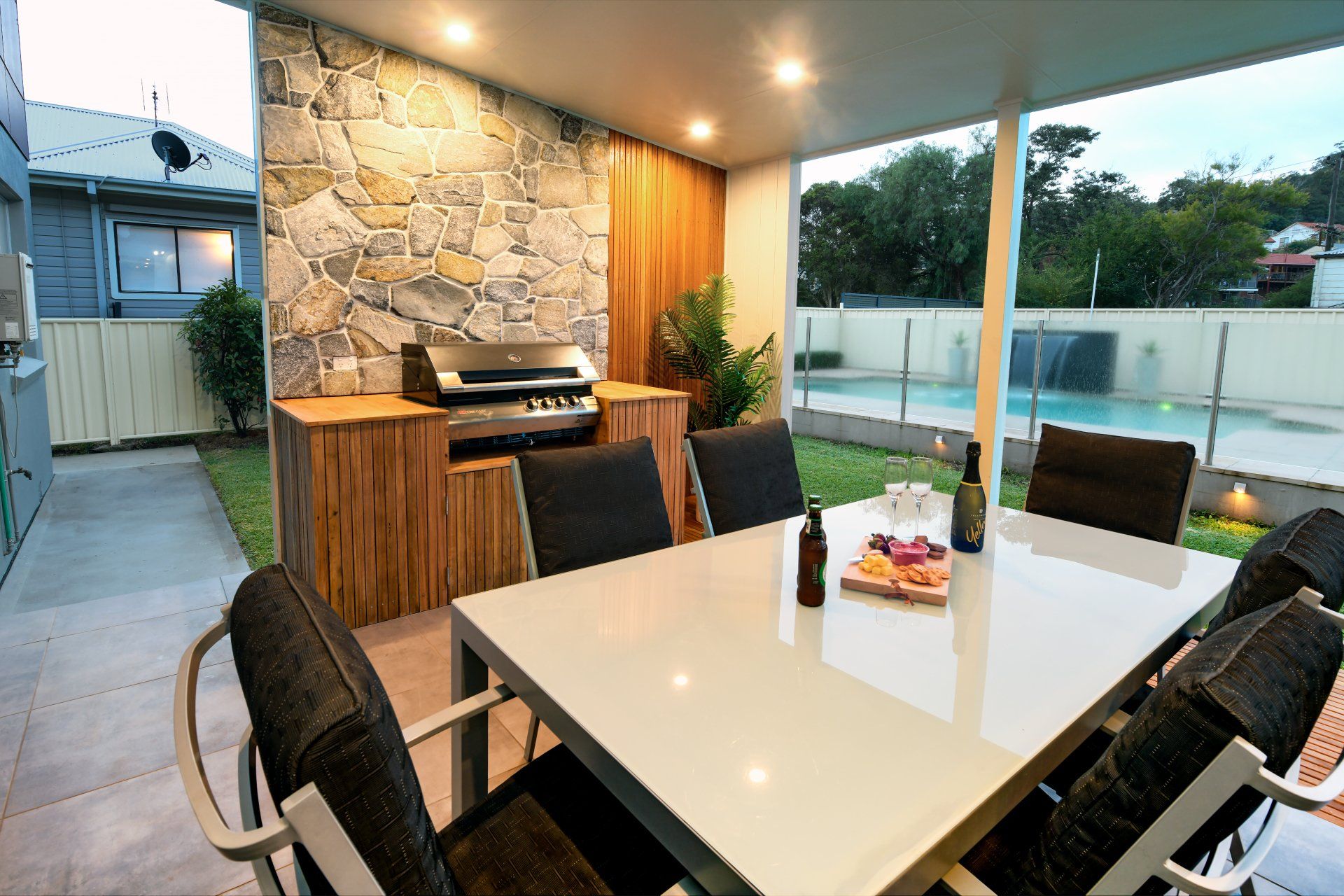 Patio with stone wall, grill, dining table with food/drinks, pool visible in the background.