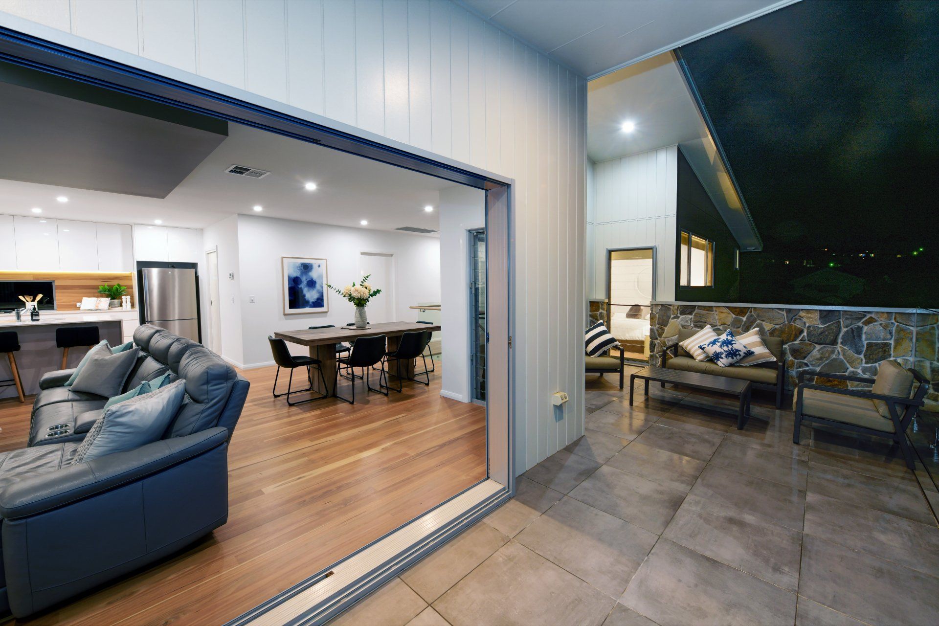 Open sliding glass door reveals a living room, dining area, and outdoor patio with seating.