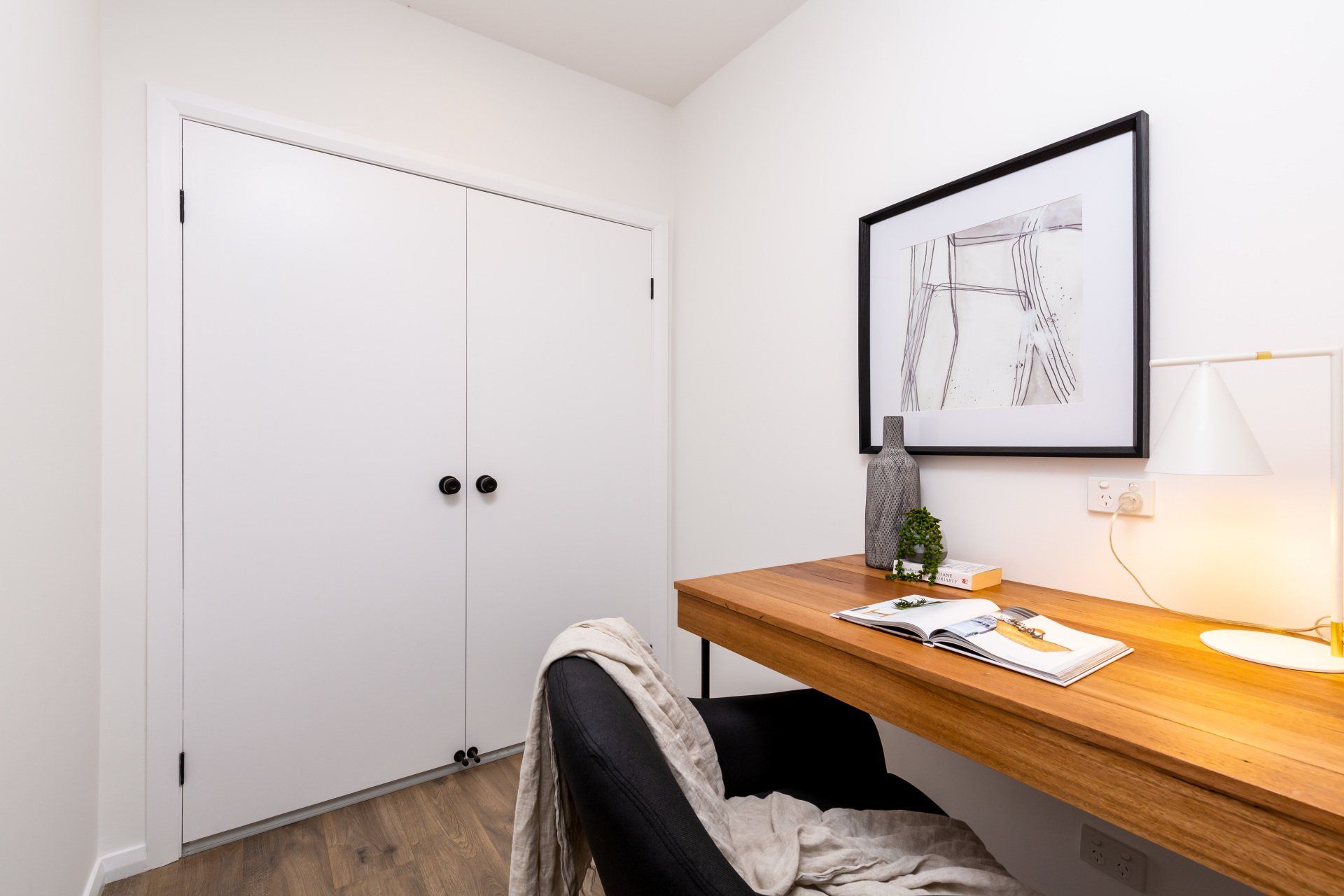 Small home office with wooden desk, black chair, and framed art. White closet doors on left.