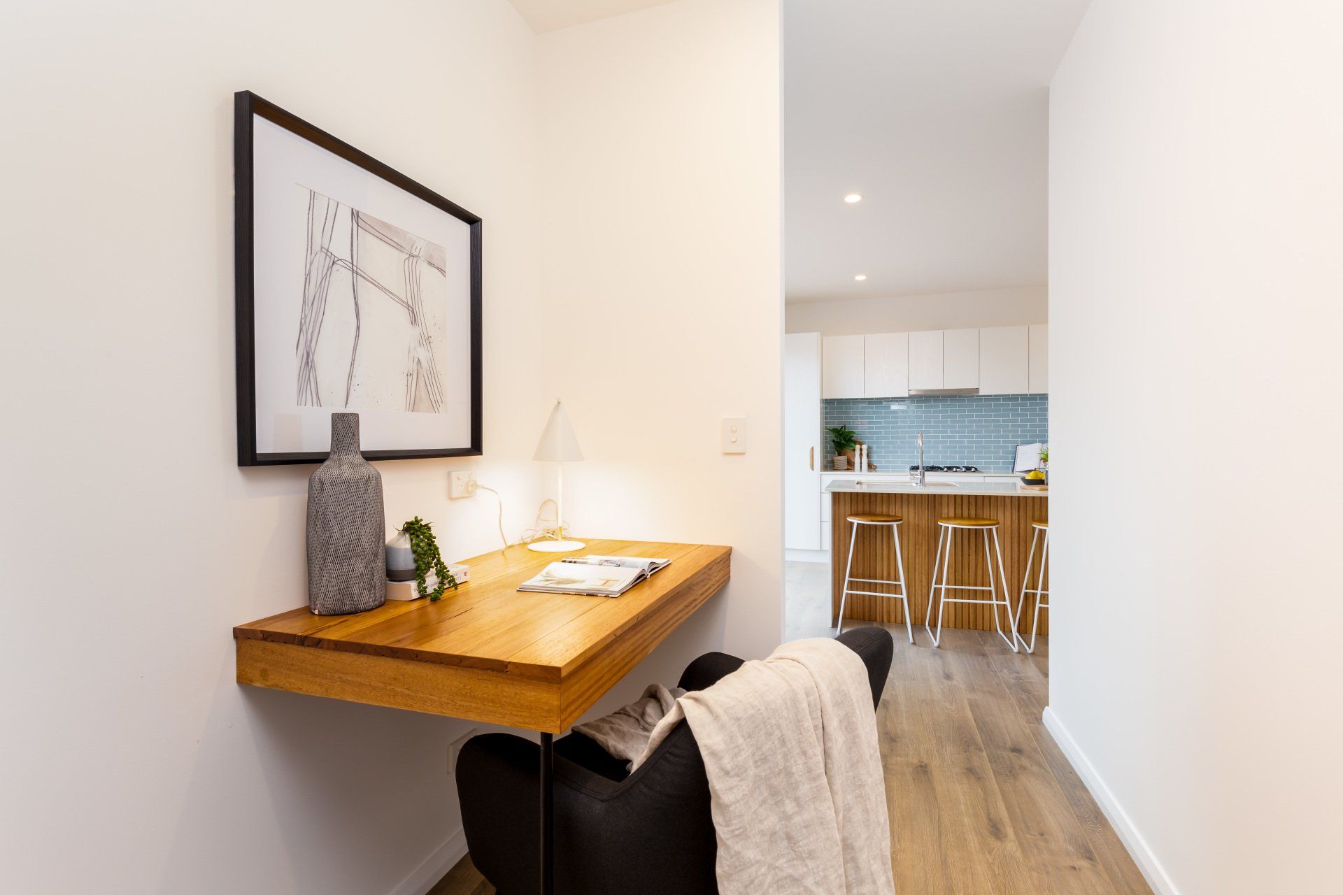 Small home office nook with a wooden desk, art, and a black chair, leading to a kitchen.